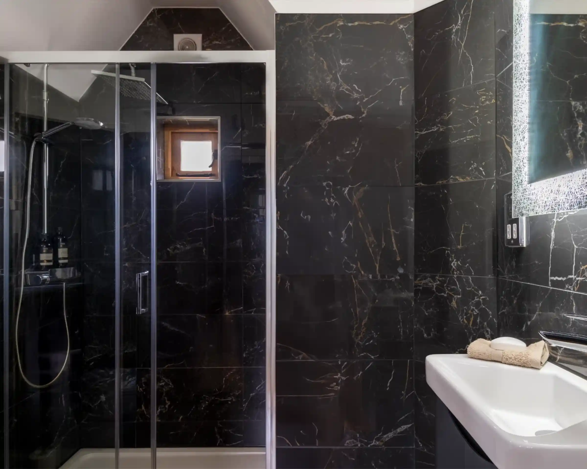 A modern bathroom featuring a glass shower enclosure, dark marble walls, and a white sink. Natural light enters through a small window, highlighting the sleek design.