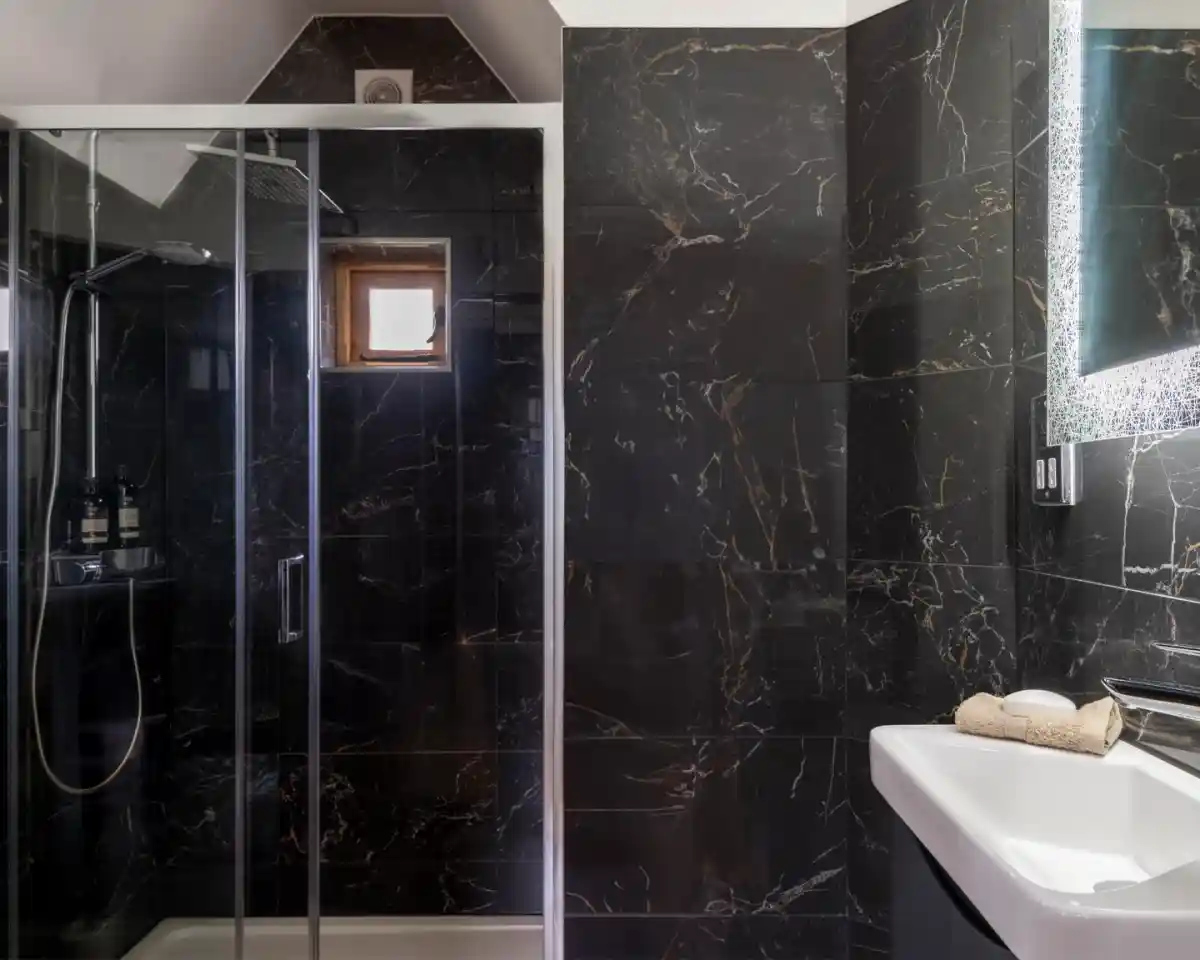 A modern bathroom featuring a glass shower enclosure, dark marble walls, and a white sink. Natural light enters through a small window, highlighting the sleek design.