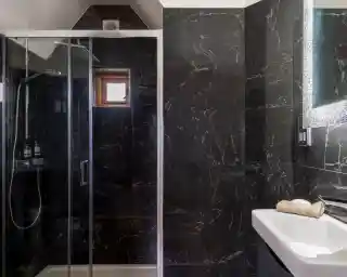 A modern bathroom featuring a glass shower enclosure, dark marble walls, and a white sink. Natural light enters through a small window, highlighting the sleek design.