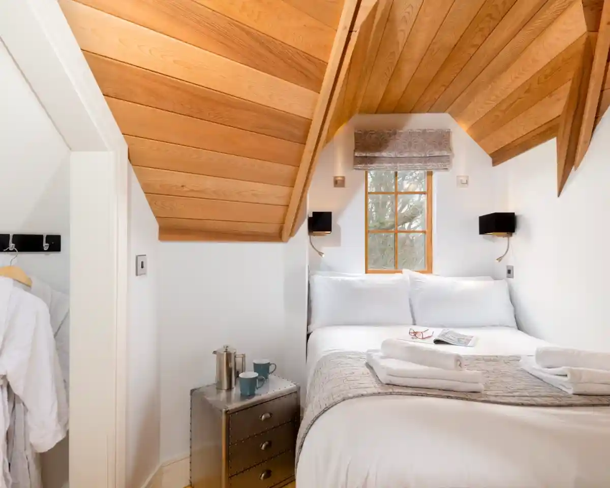 Cozy bedroom features a sloped wooden ceiling, a large bed with white linens, a bedside table with a small plant and two cups, and a reading material on the bed. Natural light comes through a window, illuminating the inviting space.