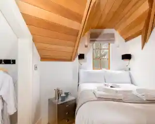 Cozy bedroom features a sloped wooden ceiling, a large bed with white linens, a bedside table with a small plant and two cups, and a reading material on the bed. Natural light comes through a window, illuminating the inviting space.