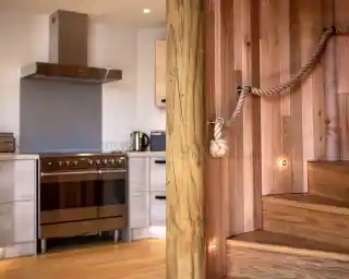 A modern kitchen with sleek cabinetry, a stainless steel oven, and a staircase made of wood with a rope handrail. The warm wooden tones and minimalist design create a cozy atmosphere.