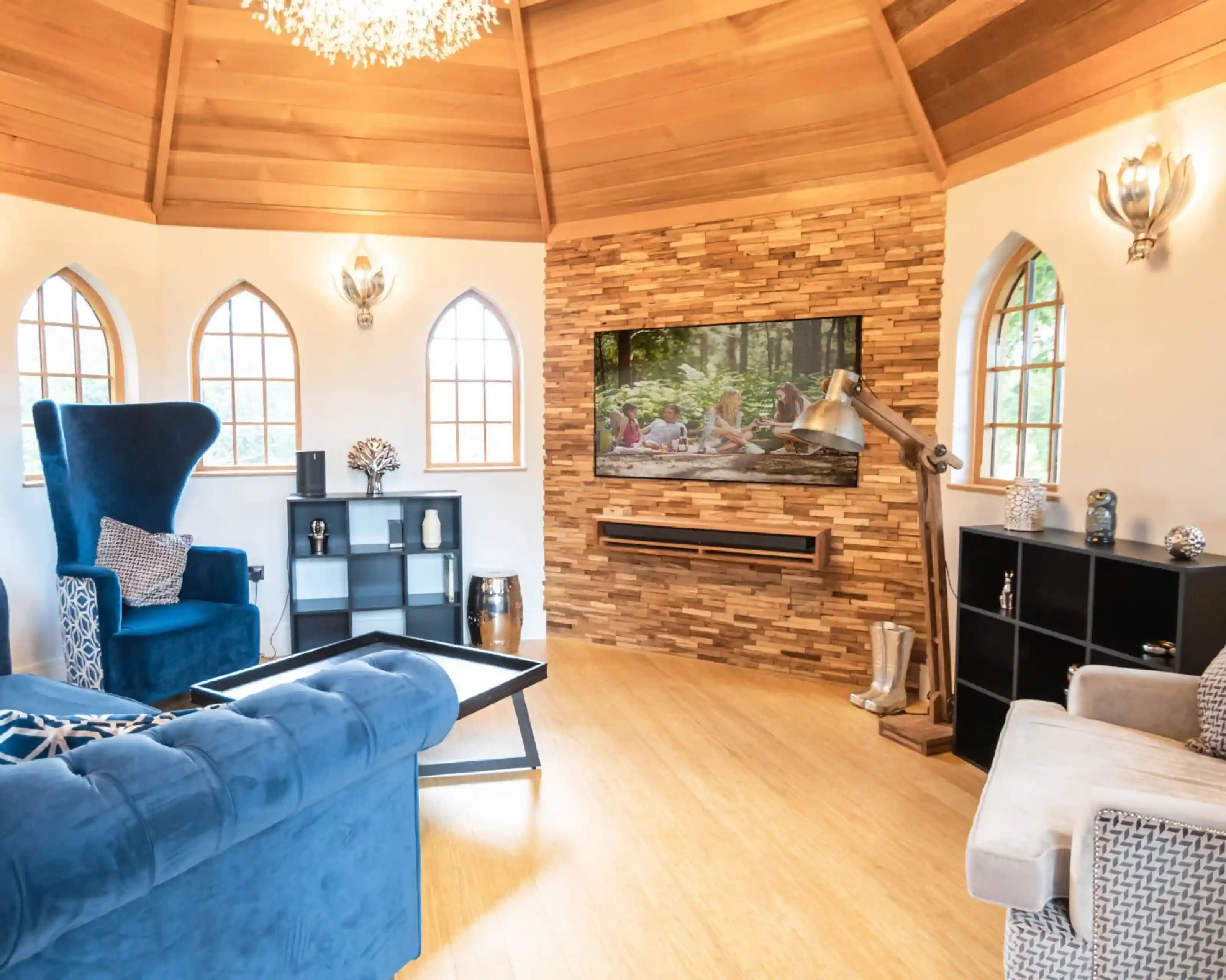 A cozy living room featuring a blue armchair and a light gray sofa. A wooden stone wall hosts a flat-screen TV above a modern wooden shelf. Large arched windows allow natural light to fill the space, accentuated by decorative lamps and accessories. A coffee table sits in the center of the room, complemented by a sleek black cube shelf.