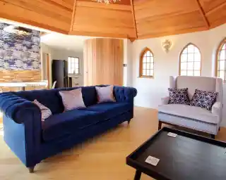 A cozy interior featuring a blue tufted sofa with decorative cushions, a light-colored armchair with patterned pillows, and a coffee table with coasters. The room has wooden ceiling beams and decorative windows, with an open kitchen in the background.