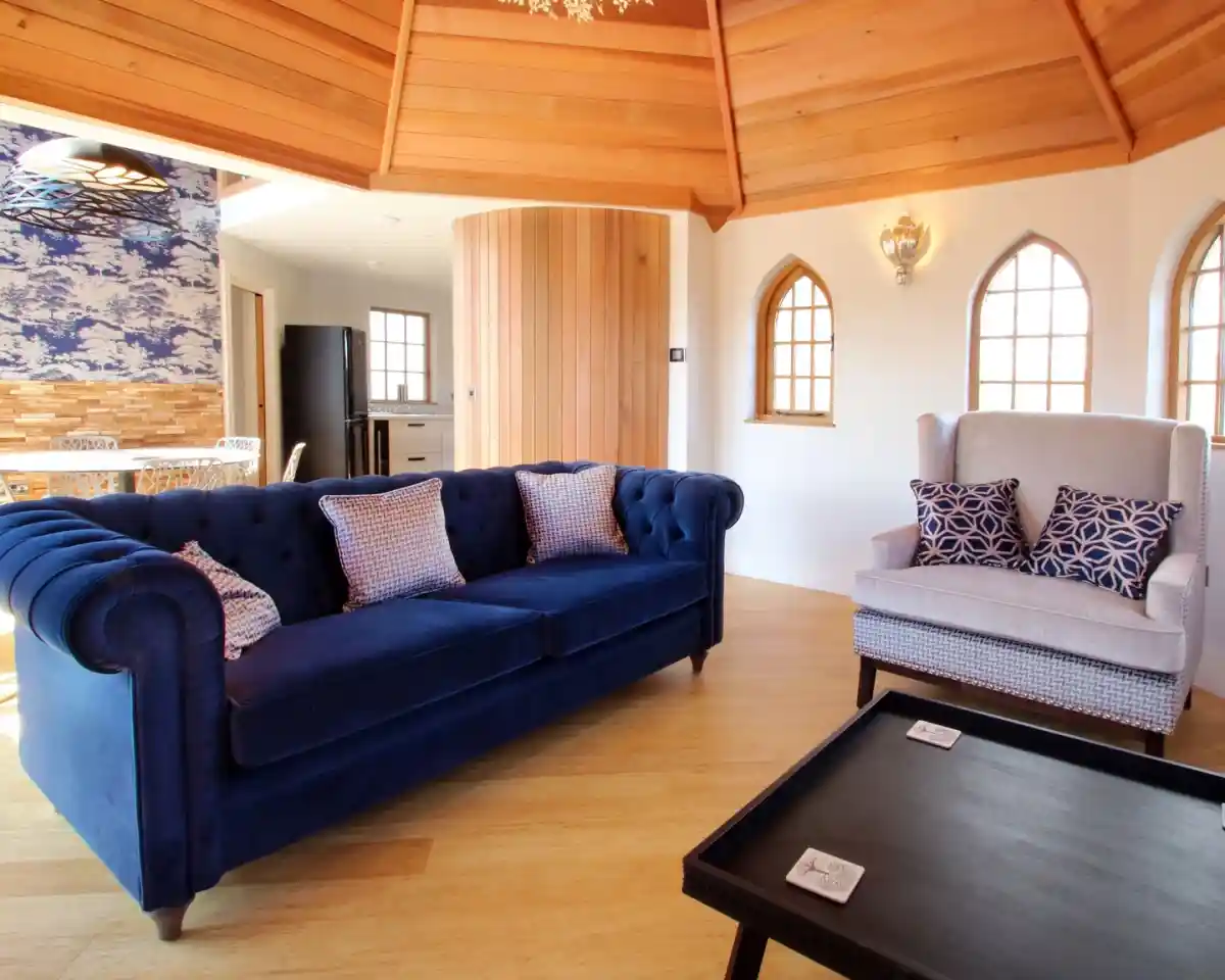 A cozy interior featuring a blue tufted sofa with decorative cushions, a light-colored armchair with patterned pillows, and a coffee table with coasters. The room has wooden ceiling beams and decorative windows, with an open kitchen in the background.