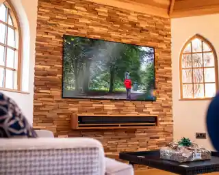A modern living room features a stone accent wall with a large flat-screen TV mounted on it. The TV displays a person in a red coat walking through a lush, green forest. In the foreground, there is a stylish coffee table with a gift-wrapped package and a cozy seating area with decorative pillows.