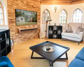 A cozy living room featuring a wooden accent wall with a flat-screen TV displaying a family outdoors. A black coffee table with a decorative tray sits in the center. In the background, a comfortable armchair and a stylish shelf can be seen, along with large windows that allow natural light to fill the space.