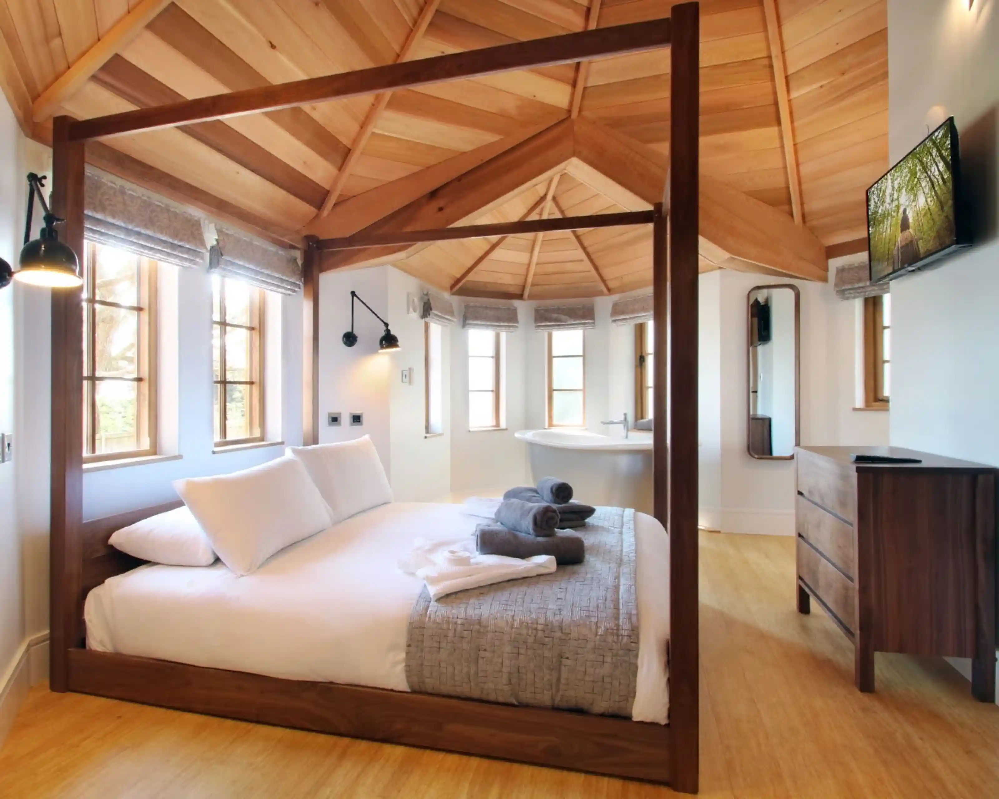A cozy bedroom featuring a four-poster bed with white linens and gray throw blankets. The room has a wooden ceiling with exposed beams, large windows allowing natural light, and modern decor including a flat-screen TV and stylish dresser.