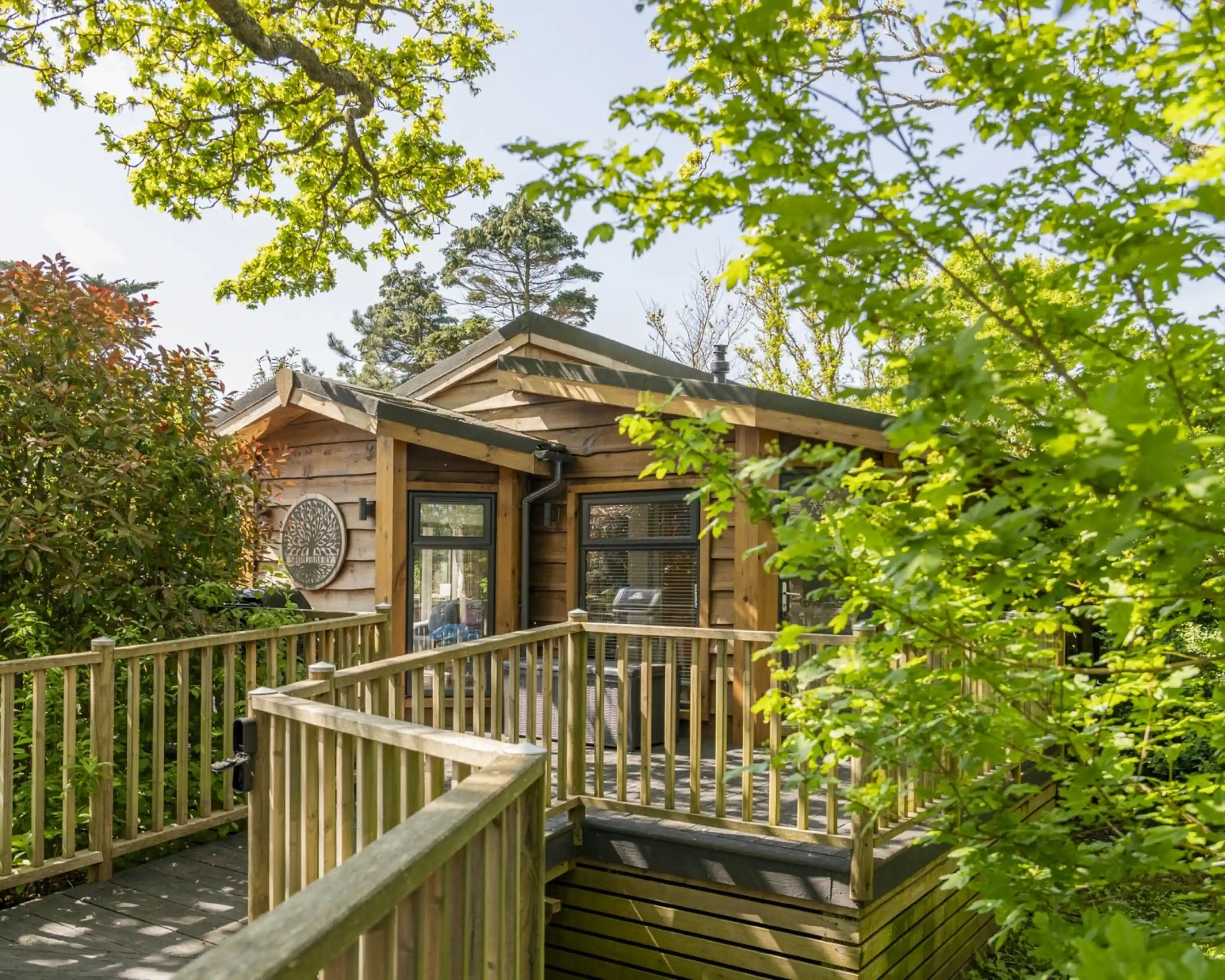 A wooden cabin surrounded by lush green foliage. A deck leads to the entrance, with a mix of trees and shrubs framing the scene against a clear blue sky.