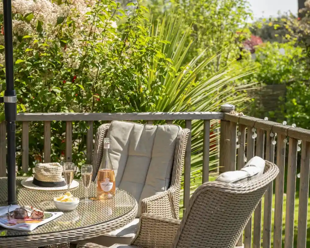 A cozy patio setting featuring a glass table with two champagne flutes, a bottle of rosé wine, and a small bowl of snacks. A sun hat rests on the table, surrounded by lush greenery and colorful flowers. Warm sunlight brightens the scene.