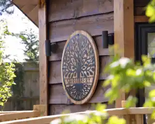 A wooden wall features an ornate circular sign with the word "Shrubbery" and a decorative tree design. Soft greenery frames the image, enhancing the natural setting.