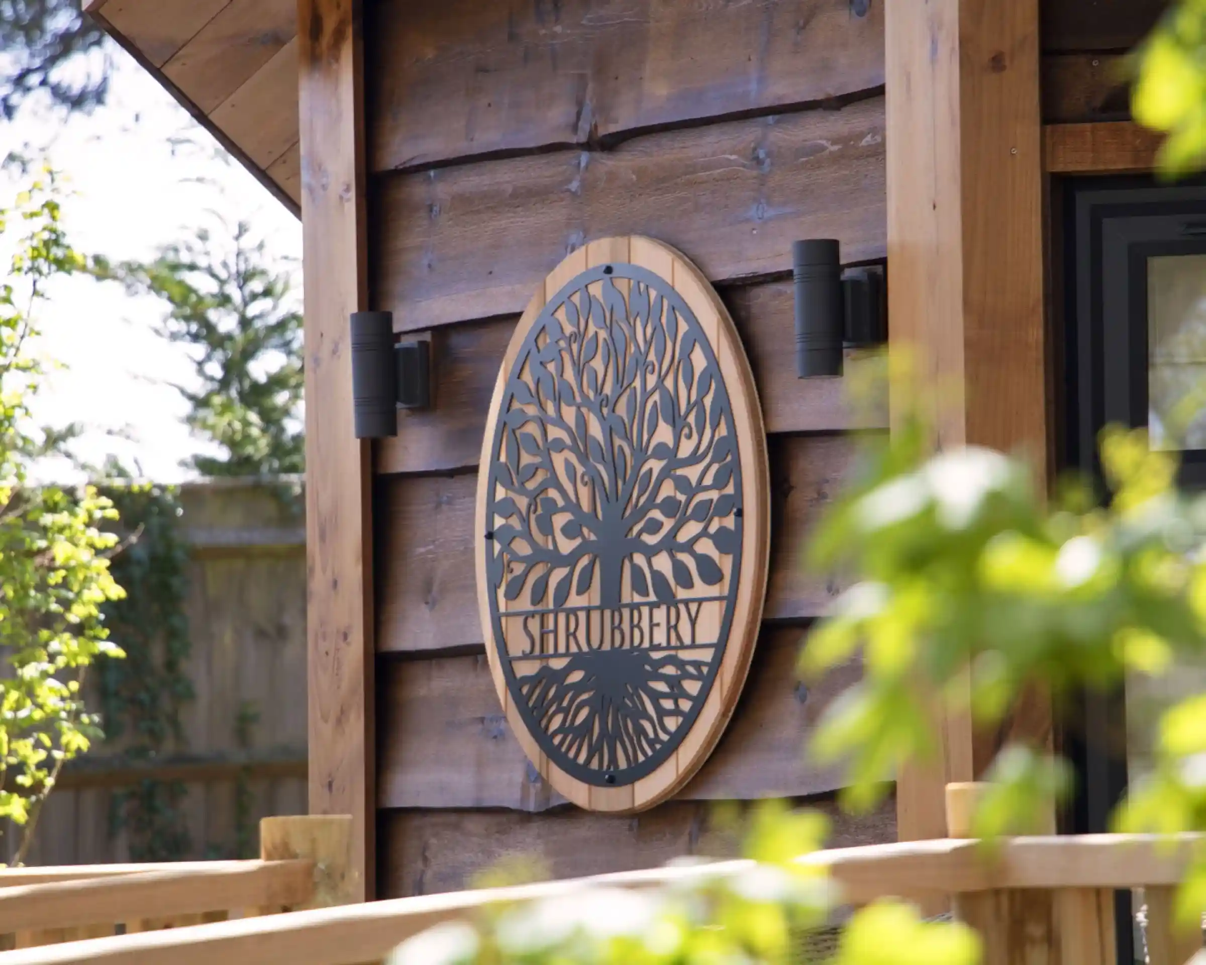 A wooden wall features an ornate circular sign with the word "Shrubbery" and a decorative tree design. Soft greenery frames the image, enhancing the natural setting.