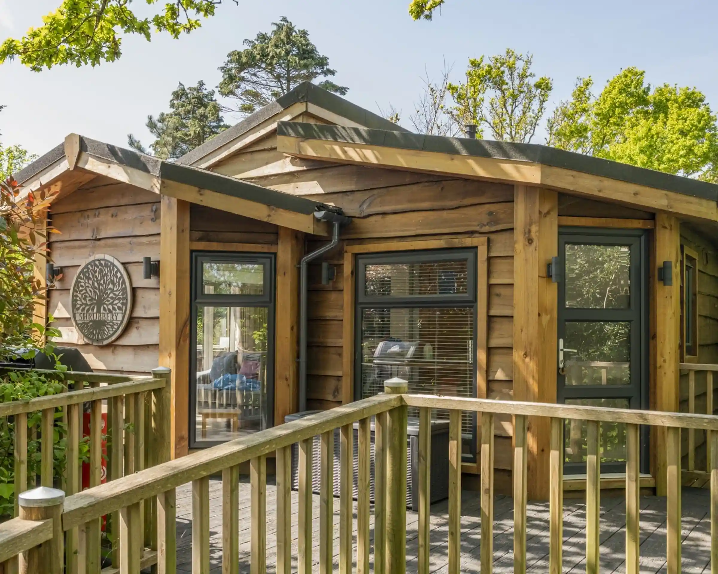 A modern wooden cabin with large windows and a rounded roof. A wooden deck leads up to the entrance, surrounded by greenery. Sunlight filters through the trees, creating a warm and inviting atmosphere.