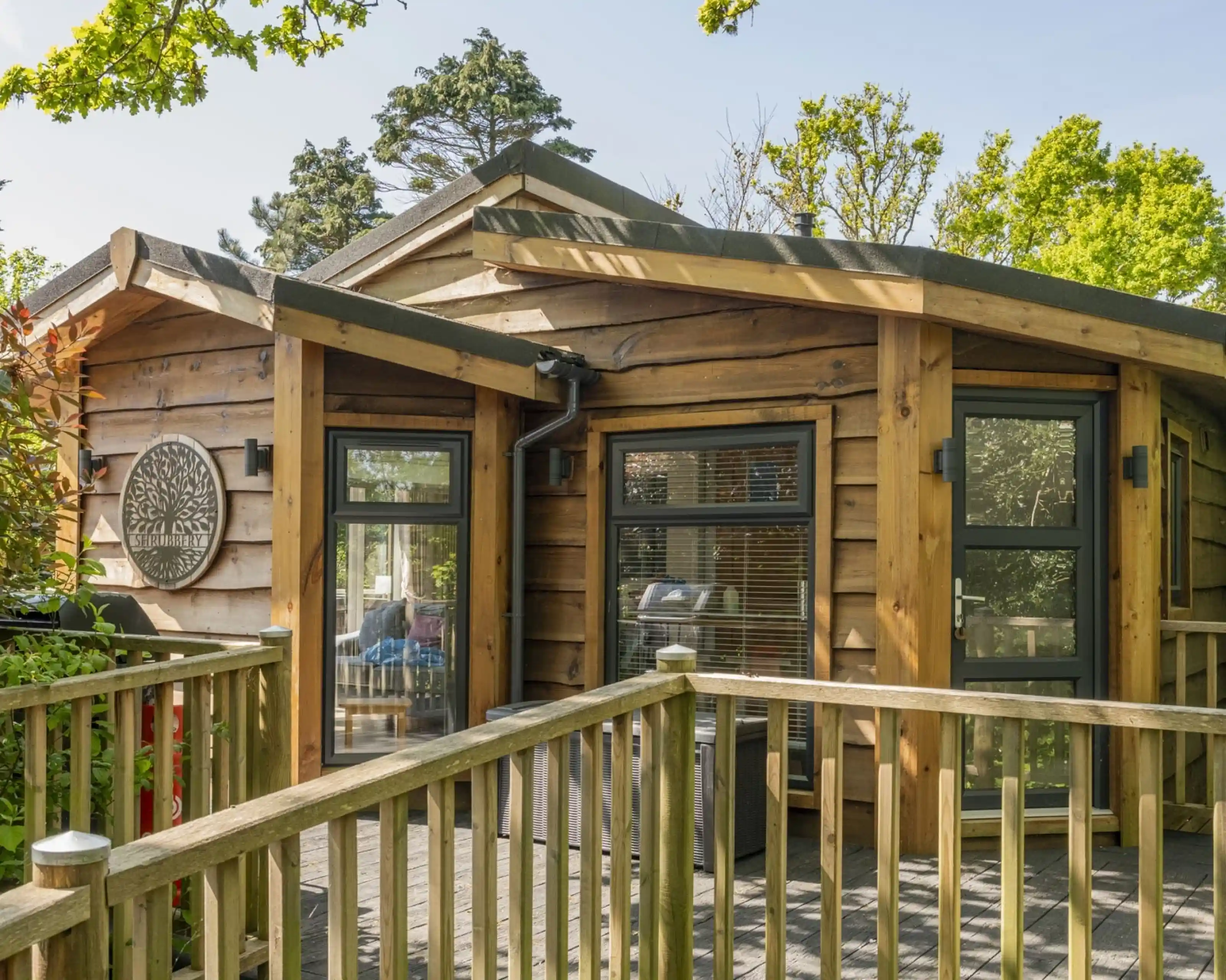 A modern wooden cabin with large windows and a rounded roof. A wooden deck leads up to the entrance, surrounded by greenery. Sunlight filters through the trees, creating a warm and inviting atmosphere.