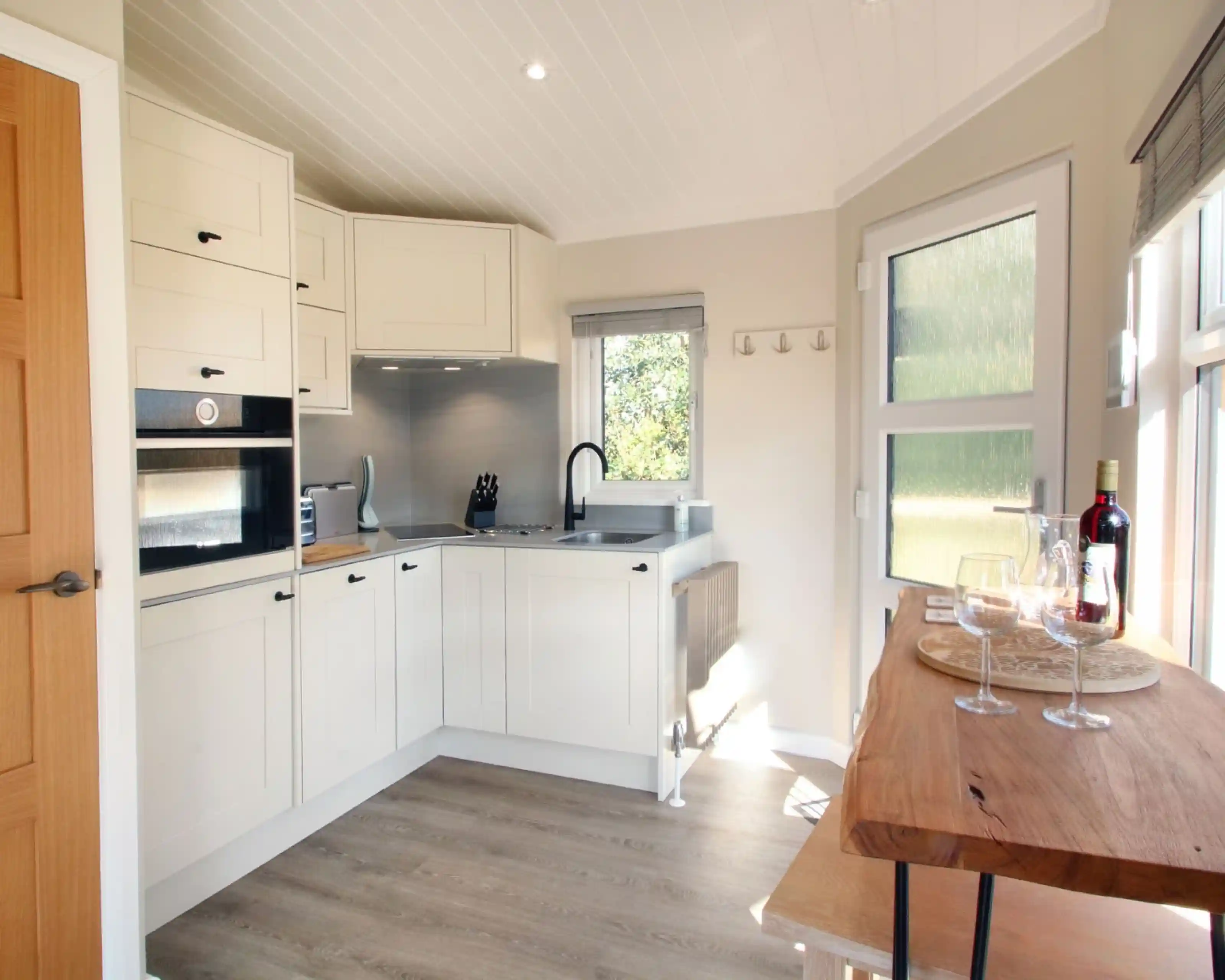 A modern kitchen featuring light-colored cabinets, a stainless steel sink, and sleek appliances. A wooden table with glasses and a bottle of wine is positioned nearby. Natural light streams in through the windows, highlighting the contemporary design.
