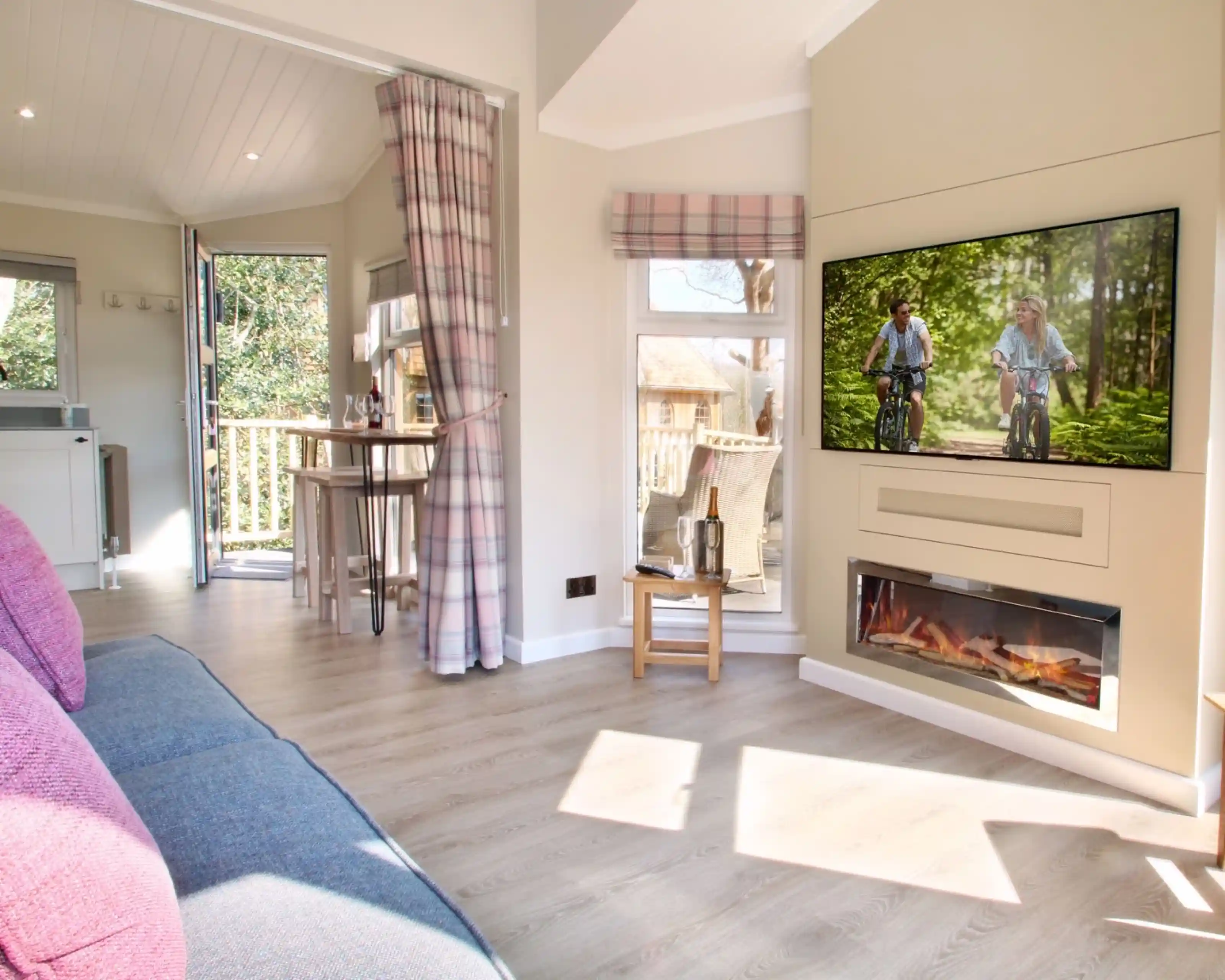 A cozy living area features a pink and gray sofa, a modern electric fireplace, and a large TV showing two children biking in a green forest. Natural light pours in from a nearby window and door leading to an outdoor deck.