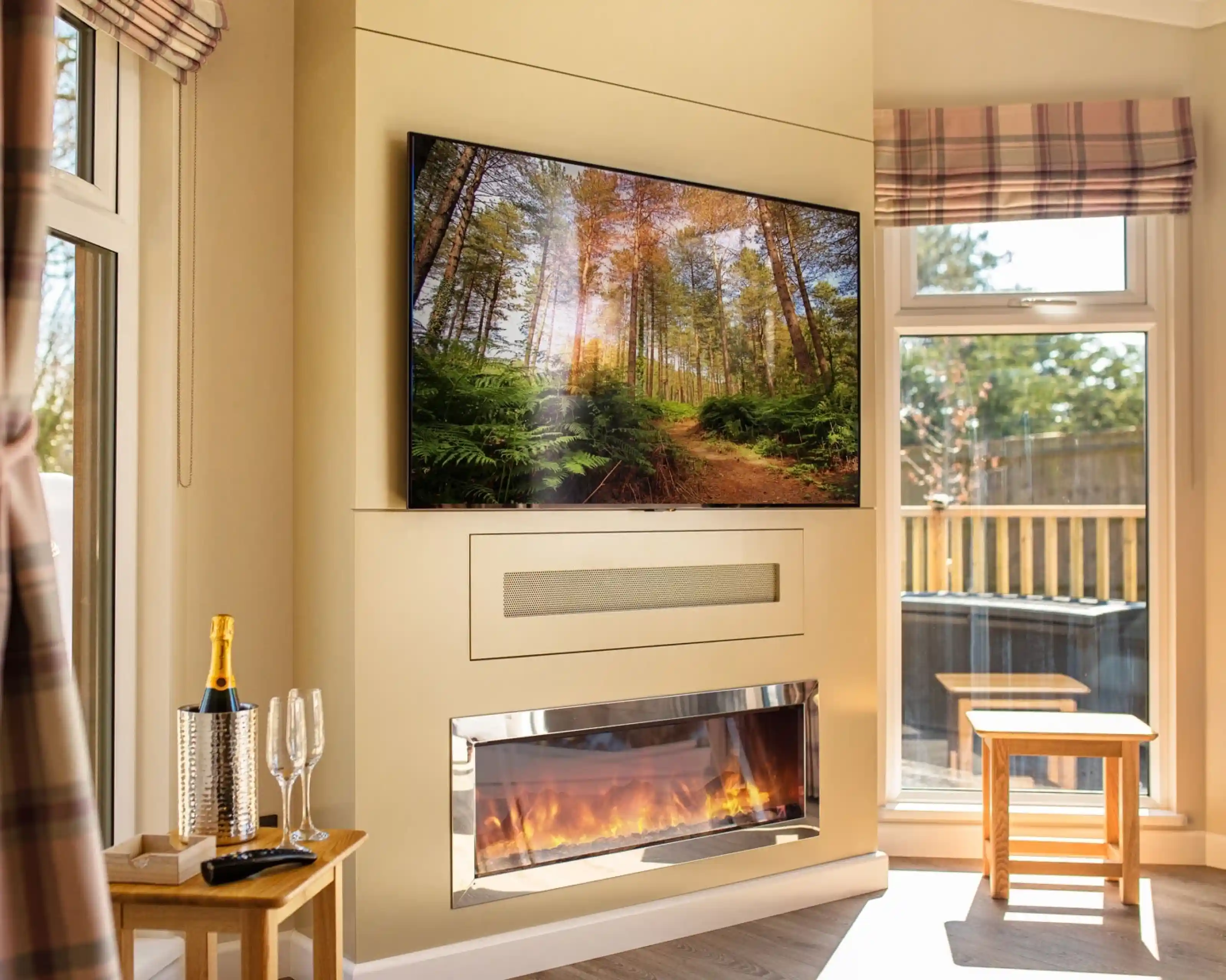 A cozy living room featuring a large television mounted on a beige wall displaying a forest scene. Below it, a sleek electric fireplace with a glowing flame adds warmth to the space. A small wooden table and two glasses are set nearby, while sunlight streams through large windows adorned with plaid valances.