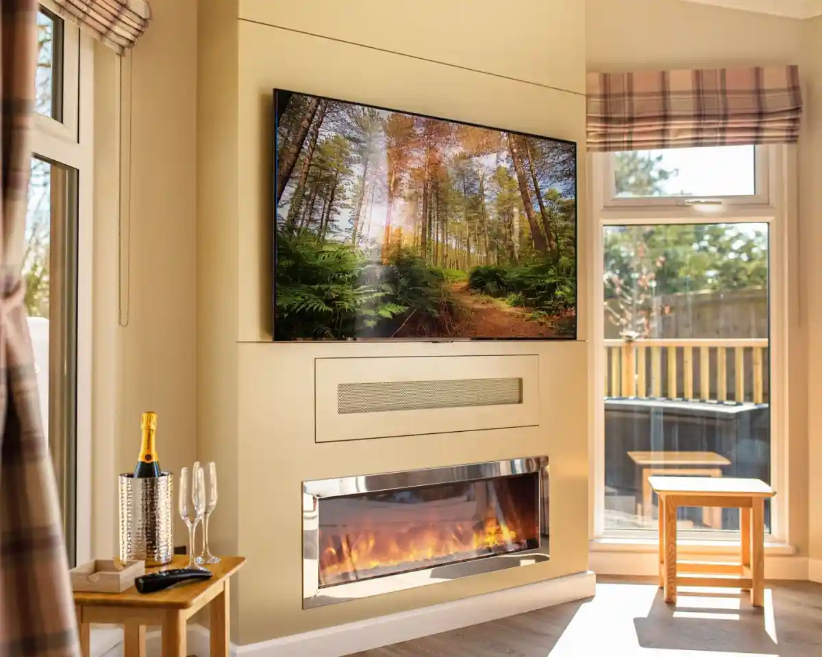A cozy living room featuring a large television mounted on a beige wall displaying a forest scene. Below it, a sleek electric fireplace with a glowing flame adds warmth to the space. A small wooden table and two glasses are set nearby, while sunlight streams through large windows adorned with plaid valances.