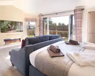 A cozy interior of a modern bedroom featuring a large bed with decorative blankets, a plush sofa, and a wall-mounted TV displaying an outdoor scene. Large sliding doors lead to a balcony, and there’s a fireplace beneath the TV, adding warmth to the space.