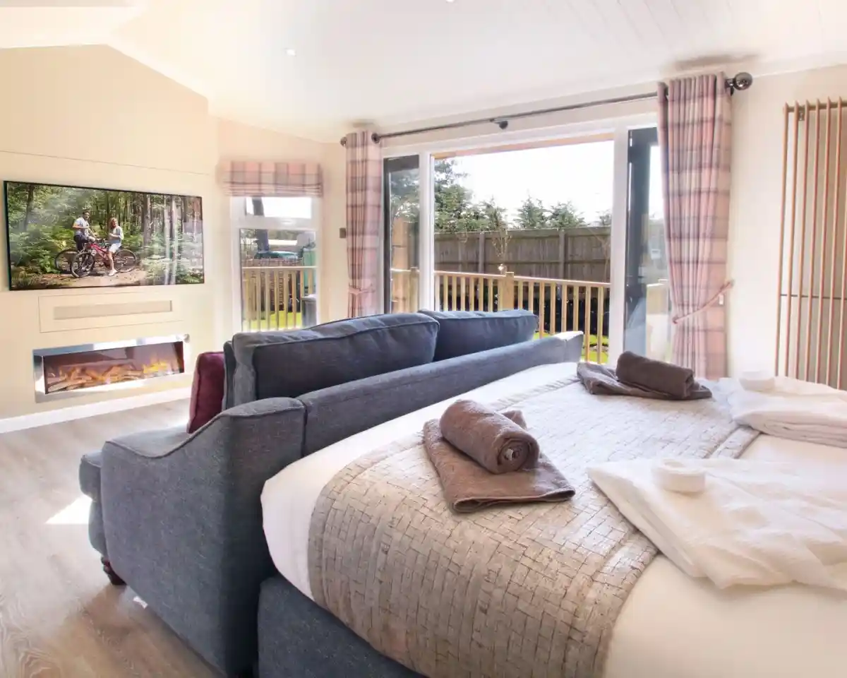A cozy interior of a modern bedroom featuring a large bed with decorative blankets, a plush sofa, and a wall-mounted TV displaying an outdoor scene. Large sliding doors lead to a balcony, and there’s a fireplace beneath the TV, adding warmth to the space.