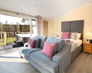 A cozy hotel room featuring a gray sofa with pink cushions, a large bed with a gray headboard, and sliding glass doors leading to a balcony. The room has light wood flooring and soft natural lighting.