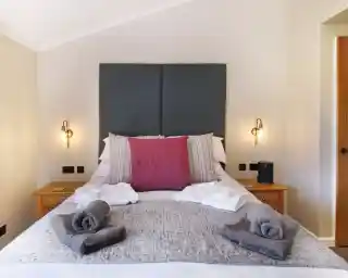 A cozy bedroom features a king-sized bed with a large, textured gray headboard. Decorative pillows, including a prominent fuchsia one, are arranged at the head of the bed. Soft, gray towels are neatly placed on the bed and nightstands. Warm lighting from wall sconces creates an inviting atmosphere. A wooden cabinet and door are visible in the background.