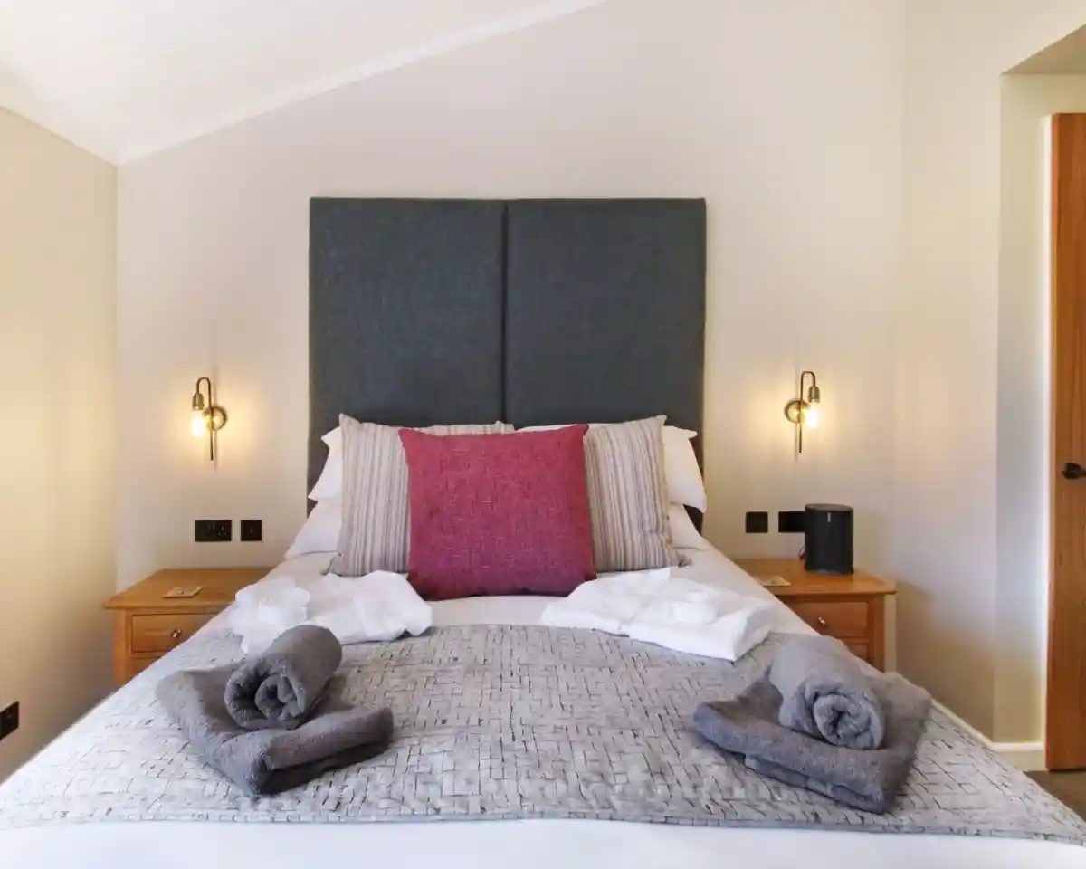 A cozy bedroom features a king-sized bed with a large, textured gray headboard. Decorative pillows, including a prominent fuchsia one, are arranged at the head of the bed. Soft, gray towels are neatly placed on the bed and nightstands. Warm lighting from wall sconces creates an inviting atmosphere. A wooden cabinet and door are visible in the background.