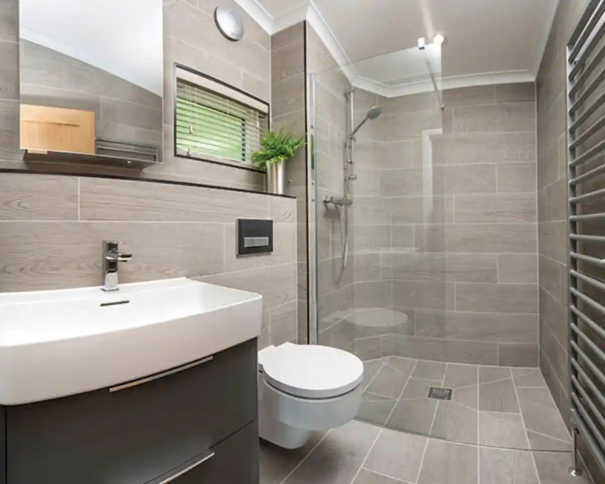 Modern bathroom featuring a glass shower enclosure, a sleek white vanity with a basin, a wall-mounted toilet, and tiled walls and floors in neutral tones. A mirror and a small plant add to the contemporary design.