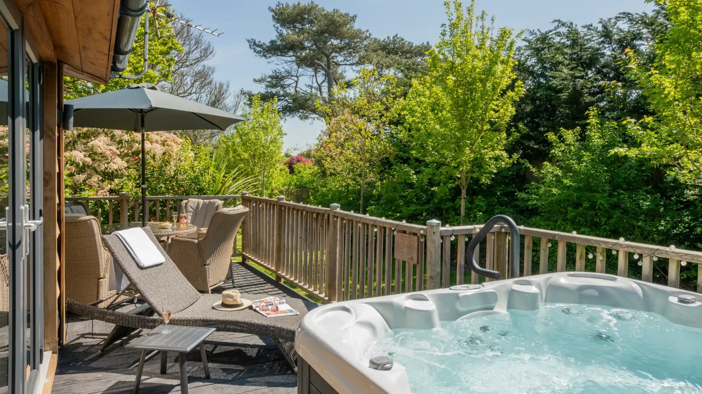 A sunny deck with a hot tub, a lounge chair, and a small table. Green trees and plants surround the area, creating a tranquil outdoor space. An umbrella provides shade.