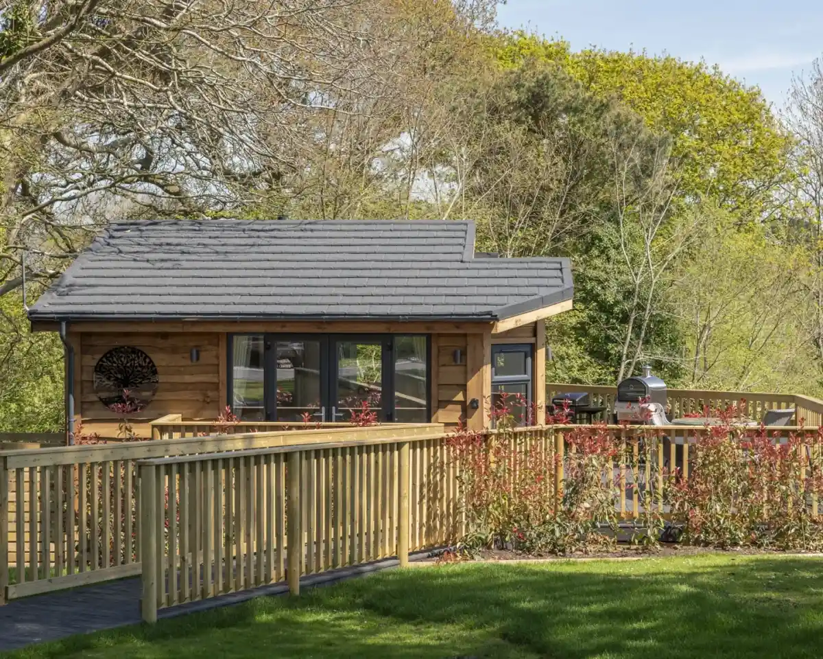 A modern wooden cabin with a gray roof is set against a backdrop of lush greenery. The cabin features large windows and a wooden deck with a railing. A flowering bush is visible in front, enhancing the serene landscape.