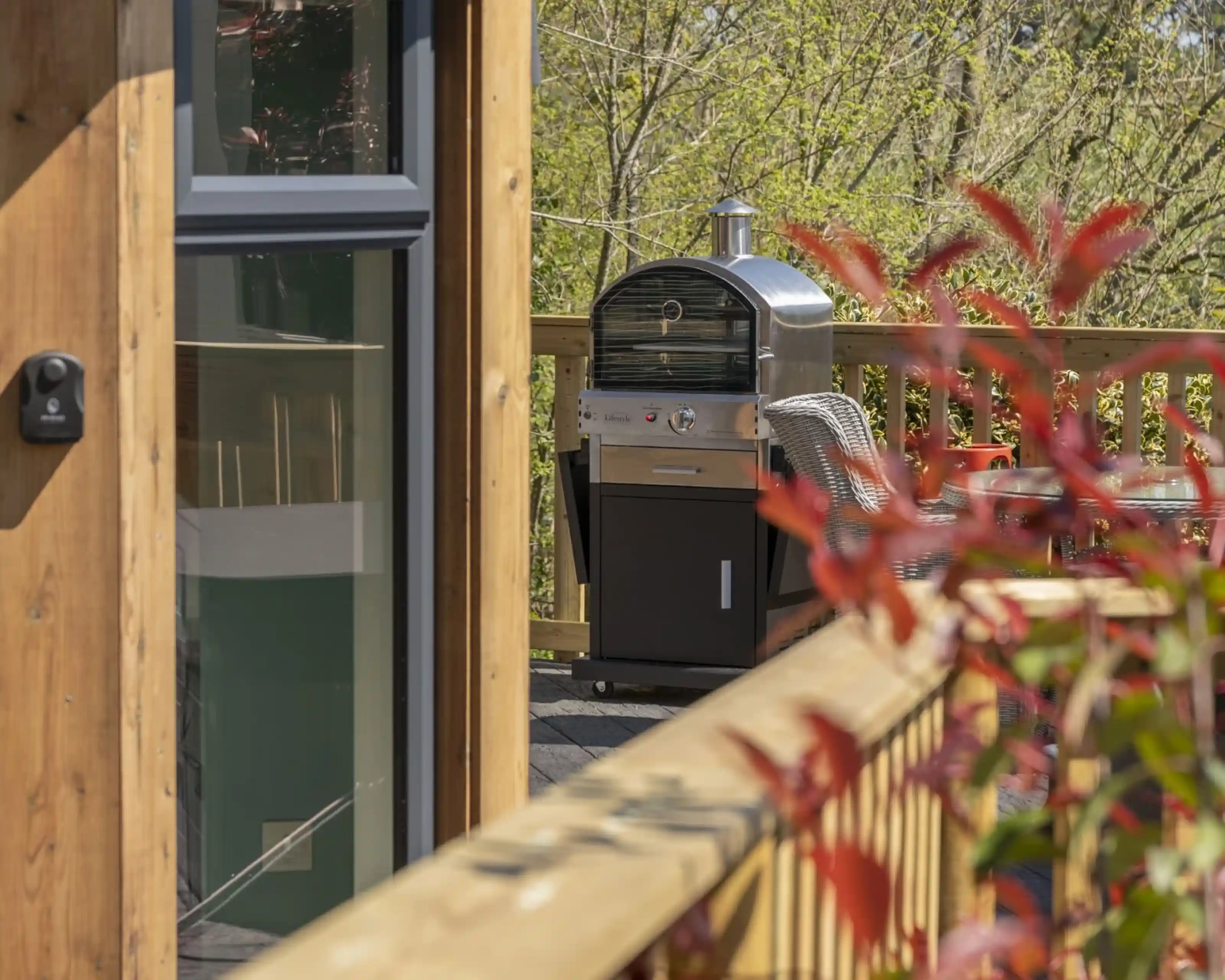 A stainless steel grill sits on a wooden deck surrounded by greenery. The deck railing is visible in the foreground, with hints of red foliage between the grill and the camera.