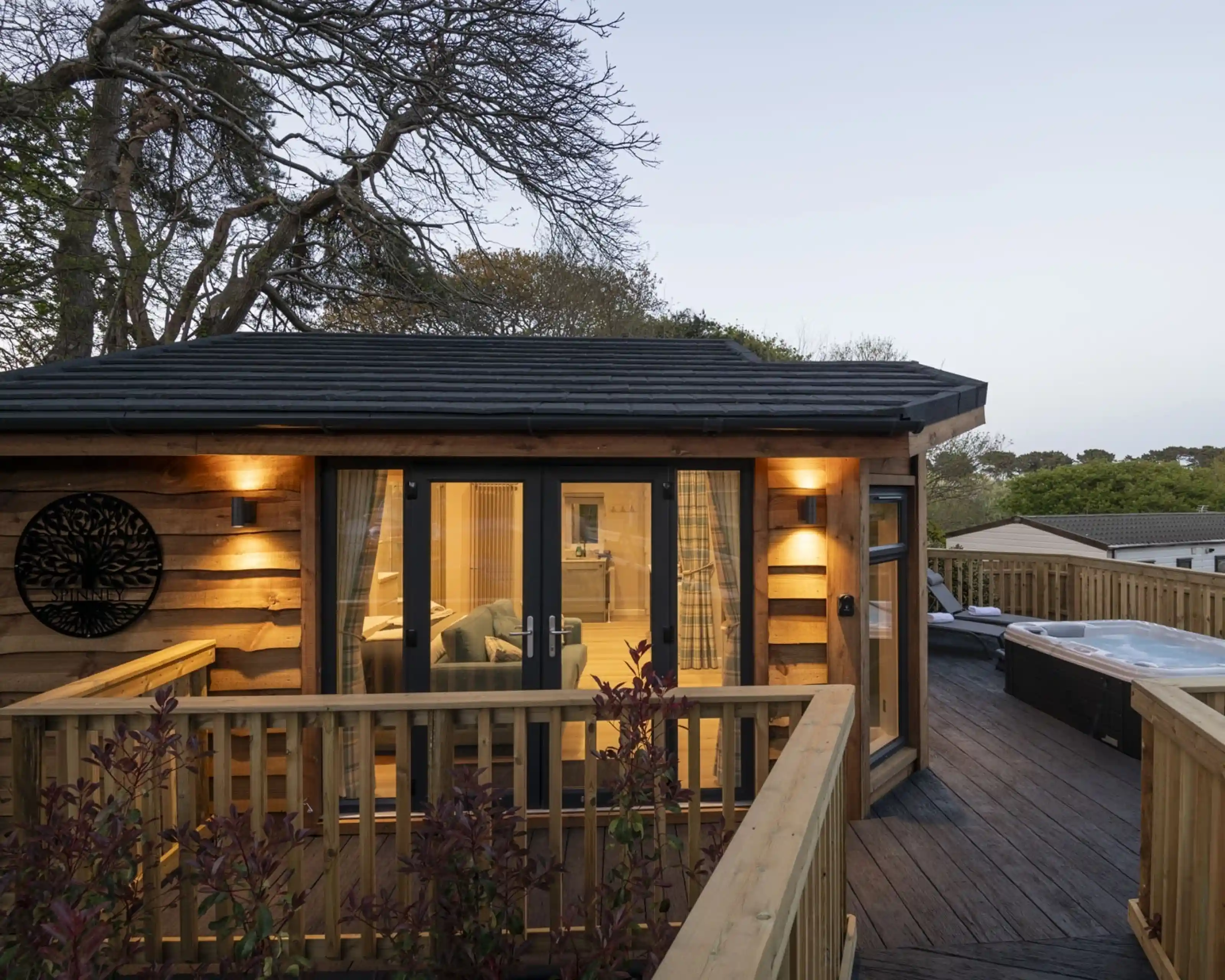 A wooden cabin with a balcony, featuring soft lighting and large glass doors. Nearby, a hot tub is visible on the deck. Surrounding trees add a natural ambiance, with twilight lighting the scene.