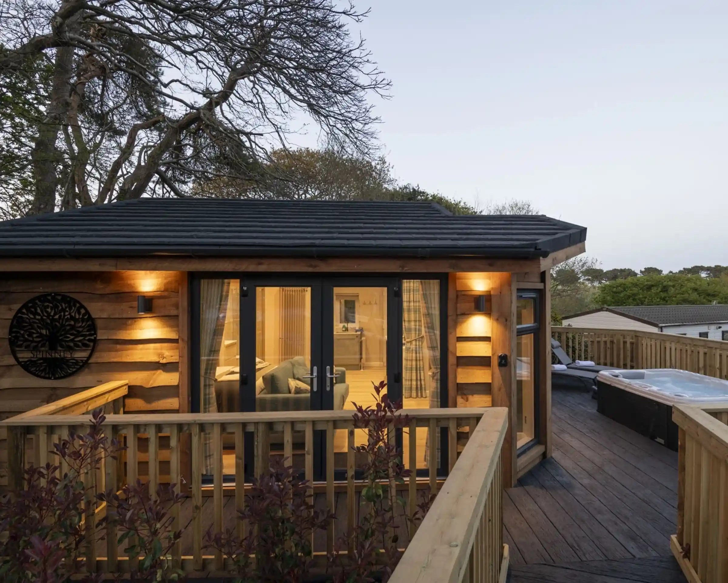 A wooden cabin with a balcony, featuring soft lighting and large glass doors. Nearby, a hot tub is visible on the deck. Surrounding trees add a natural ambiance, with twilight lighting the scene.