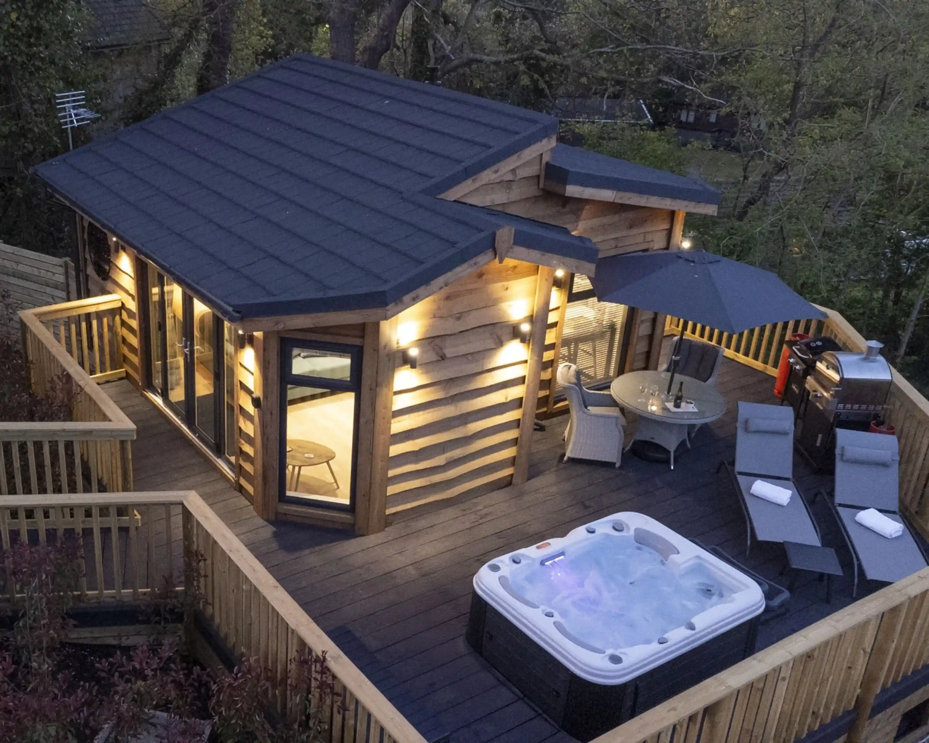 A modern wooden cabin with a sloped roof surrounded by a deck, featuring a hot tub, outdoor furniture, and an umbrella. Soft lighting illuminates the exterior, and trees are visible in the background.