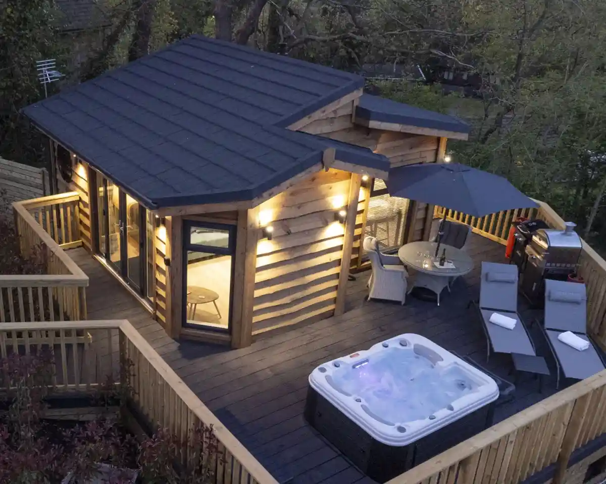 A modern wooden cabin with a sloped roof surrounded by a deck, featuring a hot tub, outdoor furniture, and an umbrella. Soft lighting illuminates the exterior, and trees are visible in the background.