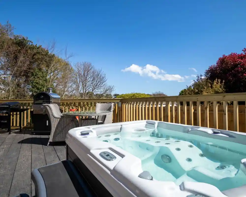 A hot tub is positioned on a wooden deck with a clear blue sky above. In the background, there are lush trees and a barbecue grill next to a stylish outdoor seating area.