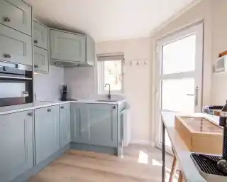 A modern kitchen with light green cabinets and marble countertops. Natural light filters through a window, highlighting the sleek appliances and minimalist design.