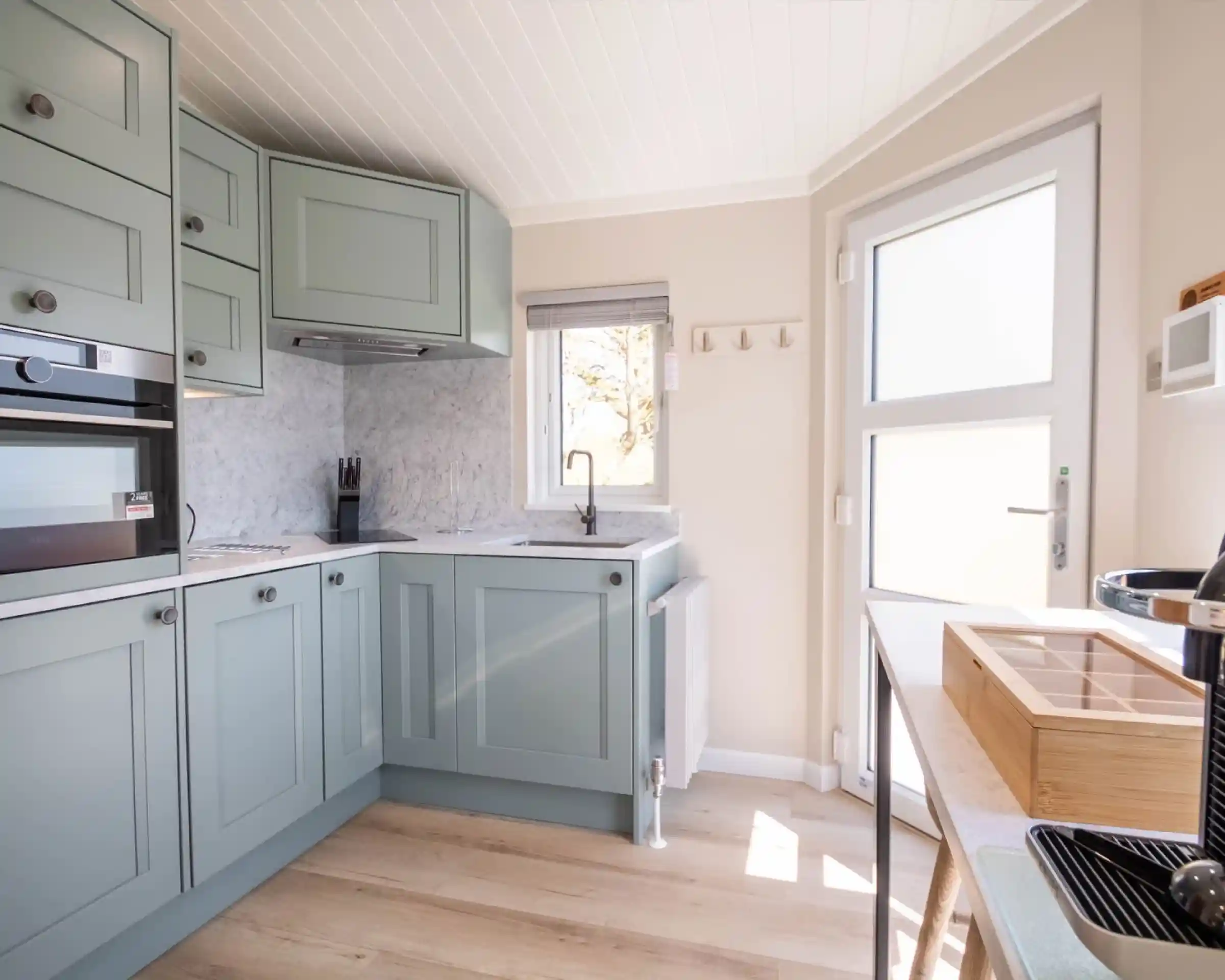 A modern kitchen with light green cabinets and marble countertops. Natural light filters through a window, highlighting the sleek appliances and minimalist design.