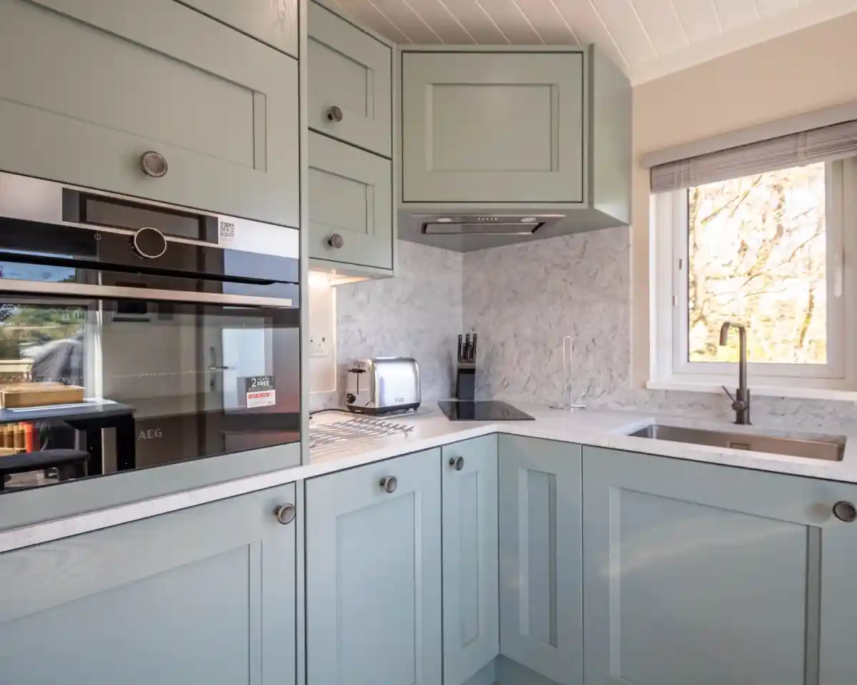 A modern kitchen featuring light green cabinets, a marble countertop, and stainless steel appliances. The layout includes a built-in oven, a sink, and a window letting in natural light.
