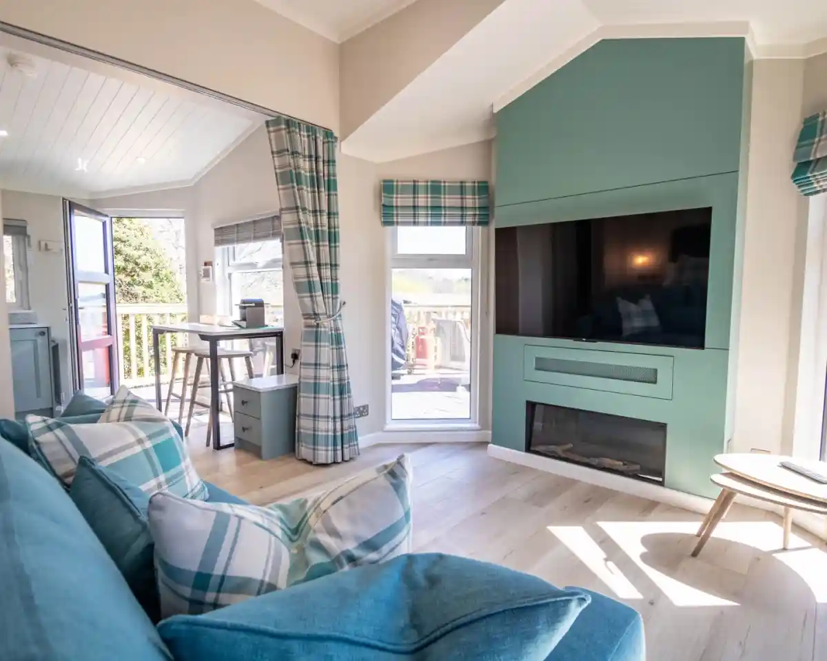 A cozy living room featuring a turquoise accent wall with a mounted television, a modern electric fireplace, and a stylish blue plaid sofa. Natural light streams through large windows, and a small dining table is visible in the background.