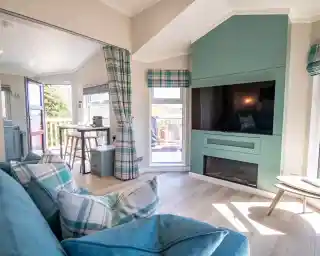 A cozy living room featuring a turquoise accent wall with a mounted television, a modern electric fireplace, and a stylish blue plaid sofa. Natural light streams through large windows, and a small dining table is visible in the background.