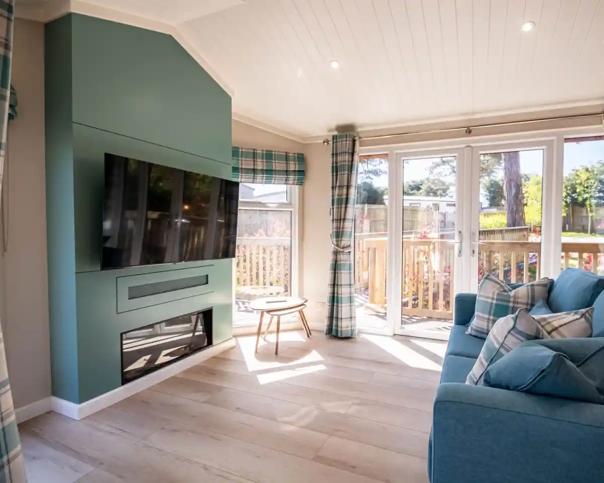 A bright and modern living room featuring a large flat-screen TV mounted on a teal accent wall, a cozy blue sofa with patterned cushions, and a small wooden table. Large glass doors open to a deck surrounded by greenery, with natural light flooding the space through windows adorned with plaid curtains.