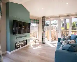 A bright and modern living room featuring a large flat-screen TV mounted on a teal accent wall, a cozy blue sofa with patterned cushions, and a small wooden table. Large glass doors open to a deck surrounded by greenery, with natural light flooding the space through windows adorned with plaid curtains.