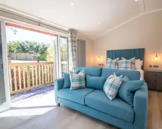A bright and modern living space features a turquoise sofa with decorative plaid pillows, large windows open to a balcony with a view of greenery outside. Soft lighting and neutral walls create a cozy atmosphere.