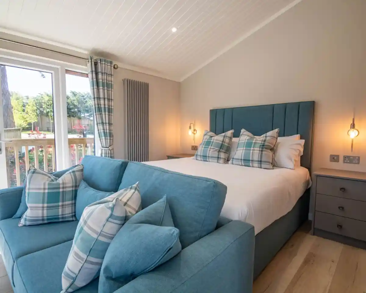 A cozy bedroom featuring a large bed with blue plaid throw pillows, a small blue sofa with matching cushions, and large windows with curtains. The room has wooden flooring and a light, airy atmosphere.