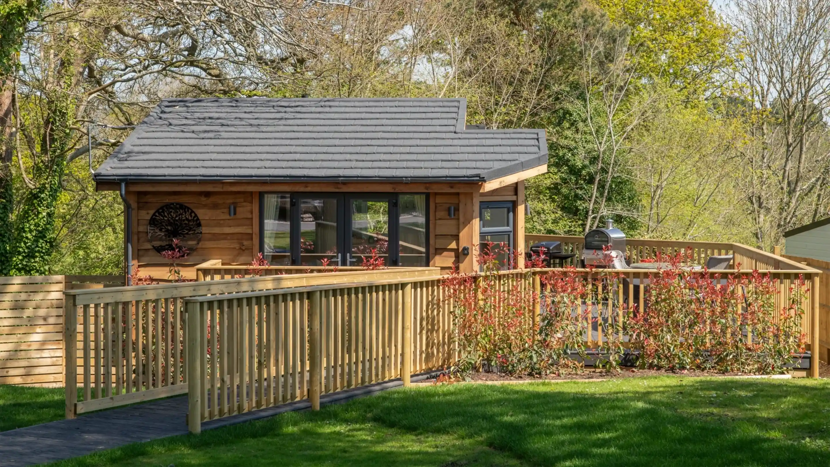 A modern wooden cabin with a sloped roof, featuring large windows and a surrounding wooden deck. The cabin is set in a lush green landscape, with blooming shrubs along the deck.