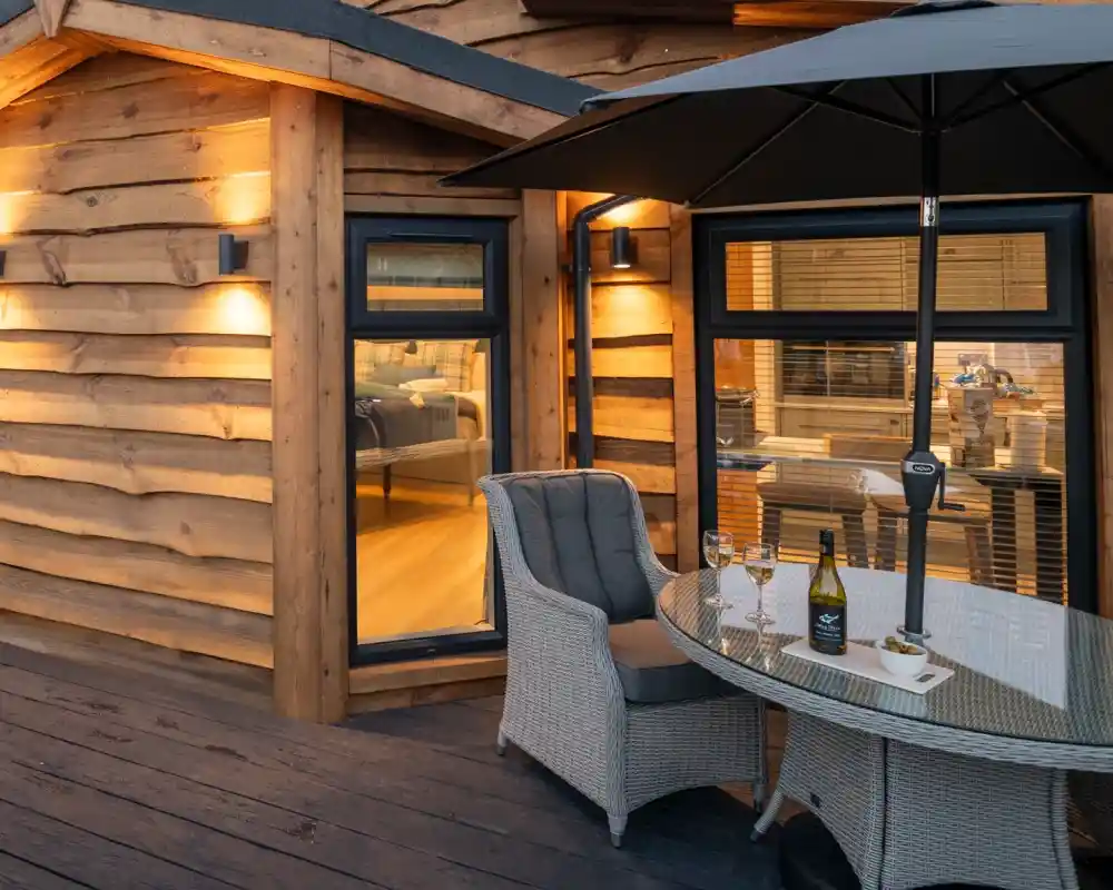 A cozy wooden cabin exterior features a small patio with a round glass table, two chairs, and an umbrella. Inside, warm light spills from a window, illuminating a comfortable interior. A bottle of wine and two glasses are set on the table.
