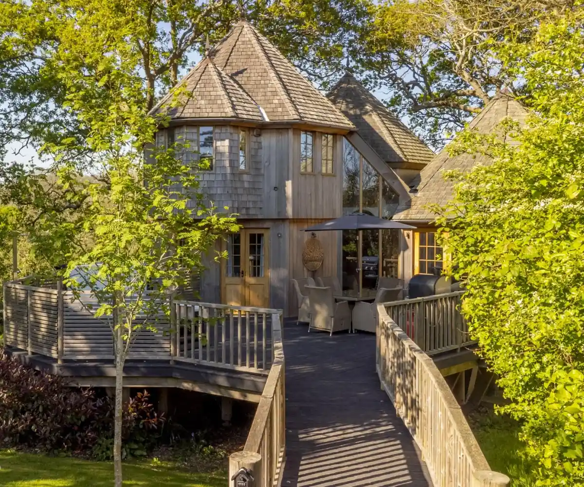 A couple stands on a balcony with a modern home in the background. The man and woman are smiling, dressed casually, with green grass and a landscaped area surrounding the house. The sky is clear and blue.