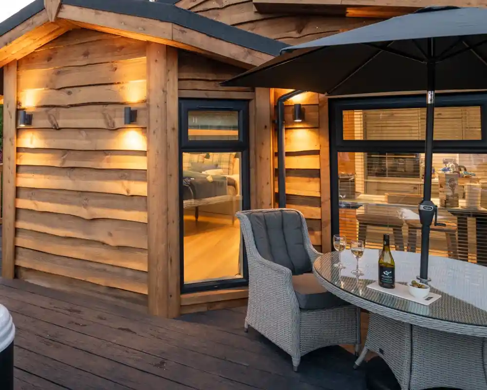 Cozy outdoor patio featuring a round table with woven chairs, a bottle of wine, and glasses, set against the backdrop of a wooden cabin with large windows and soft lighting. An umbrella provides shade for the seating area.