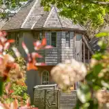 A charming wooden house with a unique turret design, surrounded by lush greenery and vibrant foliage. The structure features a shingled roof and large windows, blending harmoniously with its natural setting.